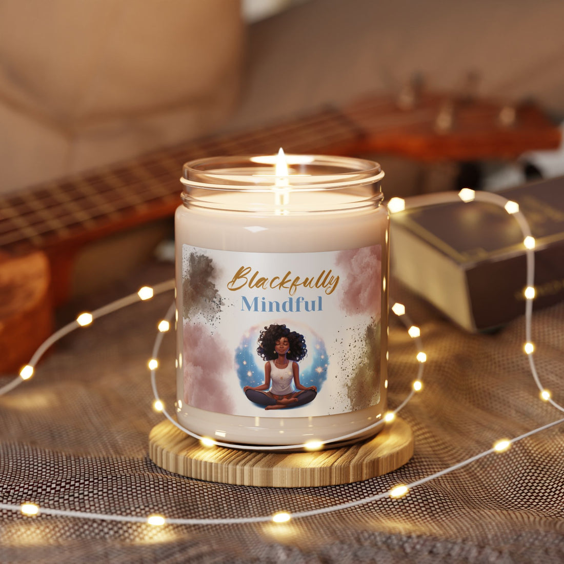 Candle in a jar labeled 'Blackfully Mindful' on a wooden coaster with fairy lights and a guitar in the background.